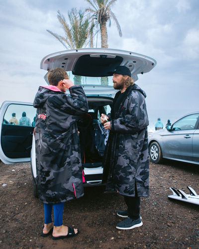 Two people in camouflage coats standing next to an open car trunk with palm trees in the background.