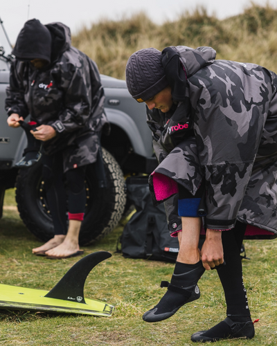 Two people in camouflage raincoats preparing for a surfing activity on grass with a vehicle in the background.