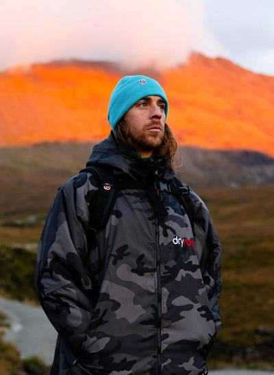 Person wearing a camouflage Dryrobe and blue beanie standing in front of a mountain with a sunset.