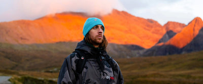 Person wearing a blue beanie and Dryrobe® standing in front of a mountain range with orange and red hues.