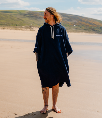 *MALE* Person wearing a navy blue hooded towel robe on a sandy beach with green hills in the background