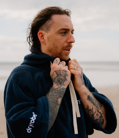 *MALE* Man wearing a navy robe with 'dryrobe' branding on a beach.
