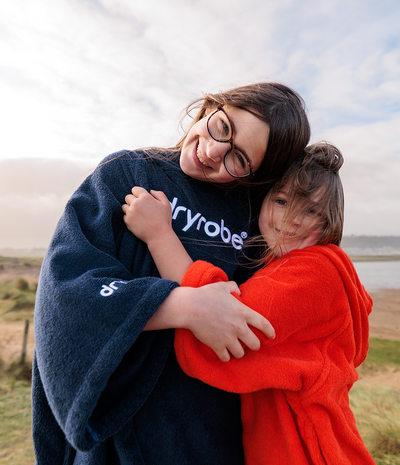 Two people hugging outdoors, one wearing a navy 'Dryrobe' towel robe and the other in a red towel robe.