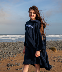 Person wearing a navy blue 'dryrobe' towel robe on a beach with ocean waves in the background