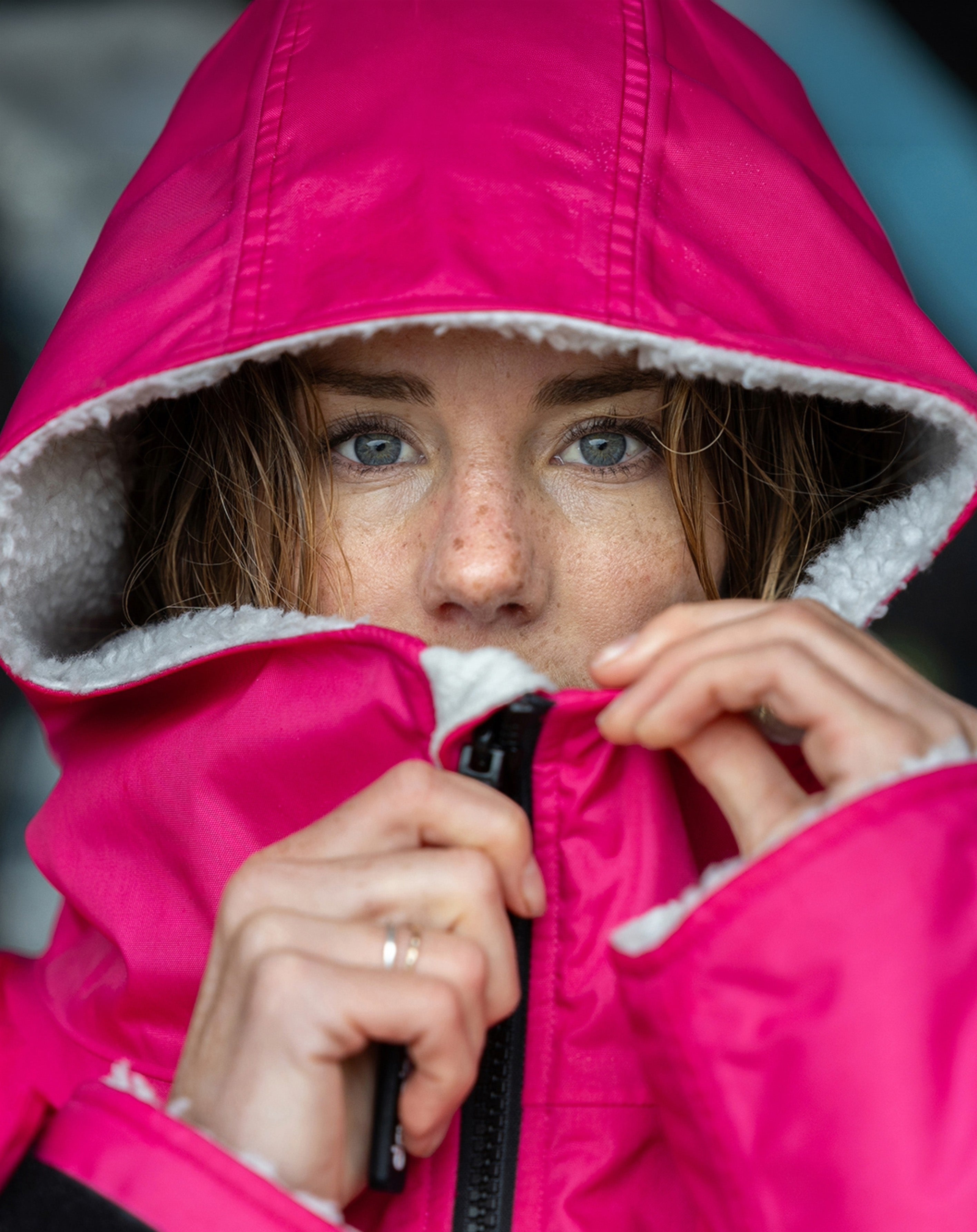 A close-up of a person wearing a bright pink hooded Dryrobe with a white sherpa lining, pulling the hood down over their face while holding a black zipper.