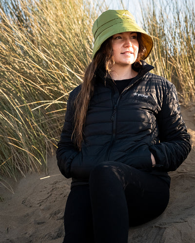 Woman sat in sand dune, wearing dryrobe Fleece Lined Bucket Hat in Forest Green and dryrobe Mid-Layer Jacket
