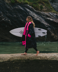 Person walking along a rocky shoreline with a surfboard, wearing a black and pink wetsuit and Dryrobe®.