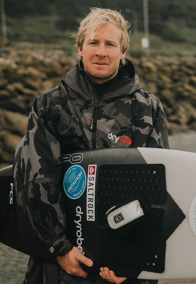 Person standing holding a surf board in a Dryrobe