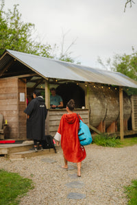 A person wrapped in a bright red blanket stands near a rustic wooden cabin outdoors, with another person visible inside the cabin. The scene includes a gravel path, greenery, and a white canopy or tent structure.