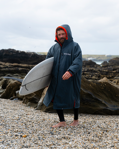 *MALE* Man holding a surfboard on a rocky beach wearing a dark poncho with red accents.