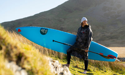 Person holding a blue paddleboard in a grassy field with mountains in the background