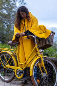 A woman wearing a bright yellow rain poncho stands beside a matching yellow bicycle with a basket, outdoors under an overcast sky.
