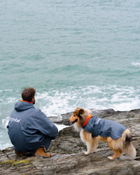 Dog wearing grey orange Dogrobe next to owner by the sea