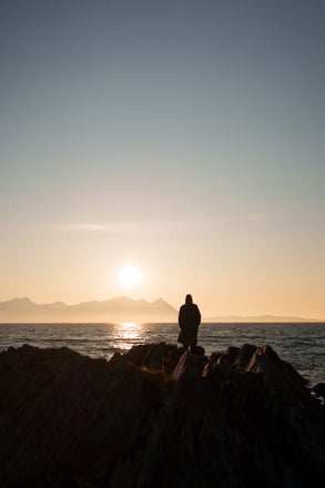 Person standing on rocks by the ocean at sunset