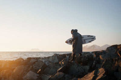 Person holding a surfboard on rocks by the ocean at sunset
