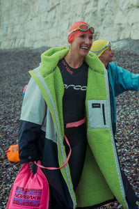 A woman on a pebble beach wearing a bright lime green fleece-lined colorblock wetsuit with a black top and orange swim cap, smiling while another person in a blue jacket stands behind her against a white chalk cliff background.
