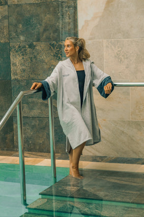 Woman in a grey Dryrobe® Towel robe standing by a poolside with marble walls.