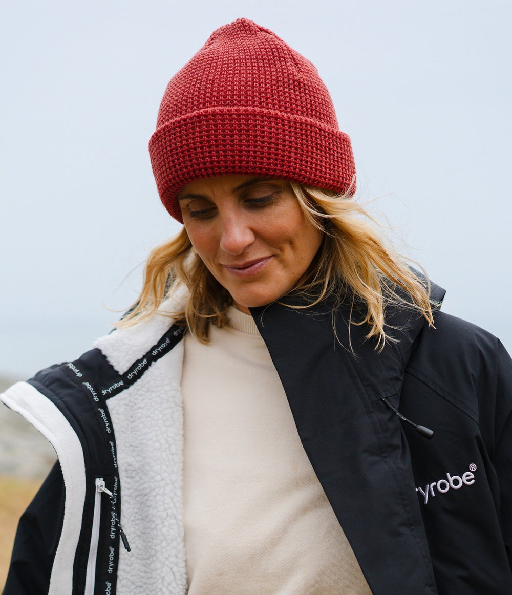A woman wearing a burnt orange knit beanie, a cream turtleneck, and a black and white Dryrobe jacket, photographed outdoors against a soft, overcast sky.