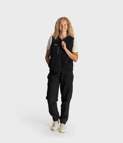 A woman modeling a black fleece vest over a cream and black short-sleeve shirt, paired with black cargo pants and beige sneakers, against a light gray studio background.