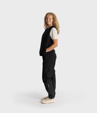 A woman stands in profile wearing a black sleeveless vest over a white short-sleeve top, black pants, and off-white sneakers against a plain light gray studio background.