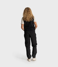 A child model shown from behind wearing a black sleeveless vest over a cream short-sleeve shirt, paired with black pants and cream sneakers, against a light gray studio background.