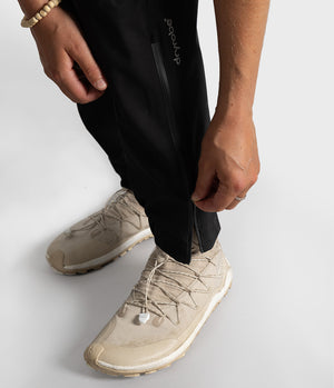Close-up of a person wearing black technical pants and beige trail running shoes, demonstrating the zippered ankle cuff of the pants.