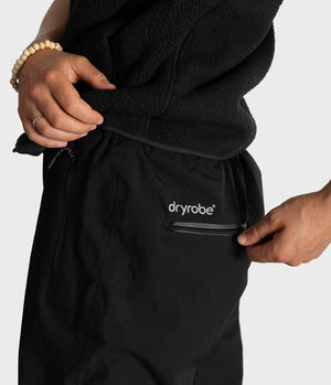 Close-up of a person wearing black Dryrobe pants, pulling up a black fleece top into the waistband, with a side zip pocket on the pants showing the white 'dryrobe' logo.