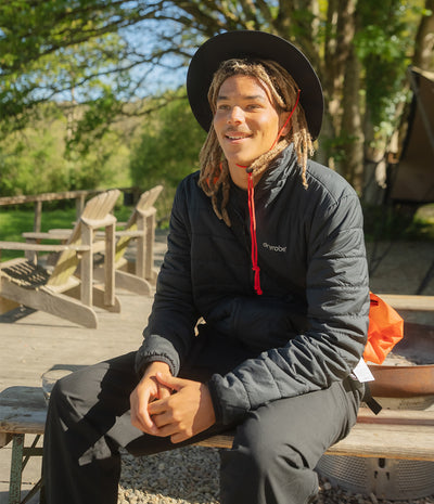 A man wearing a black wide-brim hat, a black puffer jacket with a red drawstring and white Dryrobe logo, and dark pants sits outdoors on a wooden bench in a sunny, wooded setting.