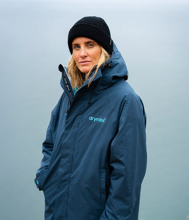A woman wearing a navy blue hooded dryrobe jacket and a black knit beanie, photographed against a soft, misty gray-blue outdoor background.
