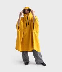 A model wearing a bright yellow hooded rain poncho over gray jeans, standing against a light gray studio background.