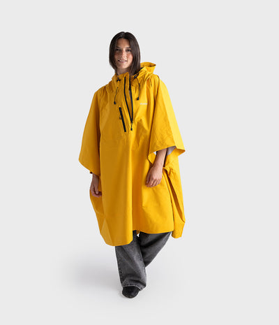 A model wearing a bright yellow hooded rain poncho with black drawstrings and zipper details, paired with gray jeans, against a light gray studio background.