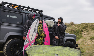 Three people with surfboards standing next to a Land Rover in a coastal setting.