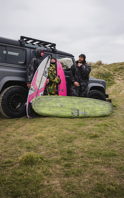 Two people with surfboards standing next to a vehicle on a grassy hillside.
