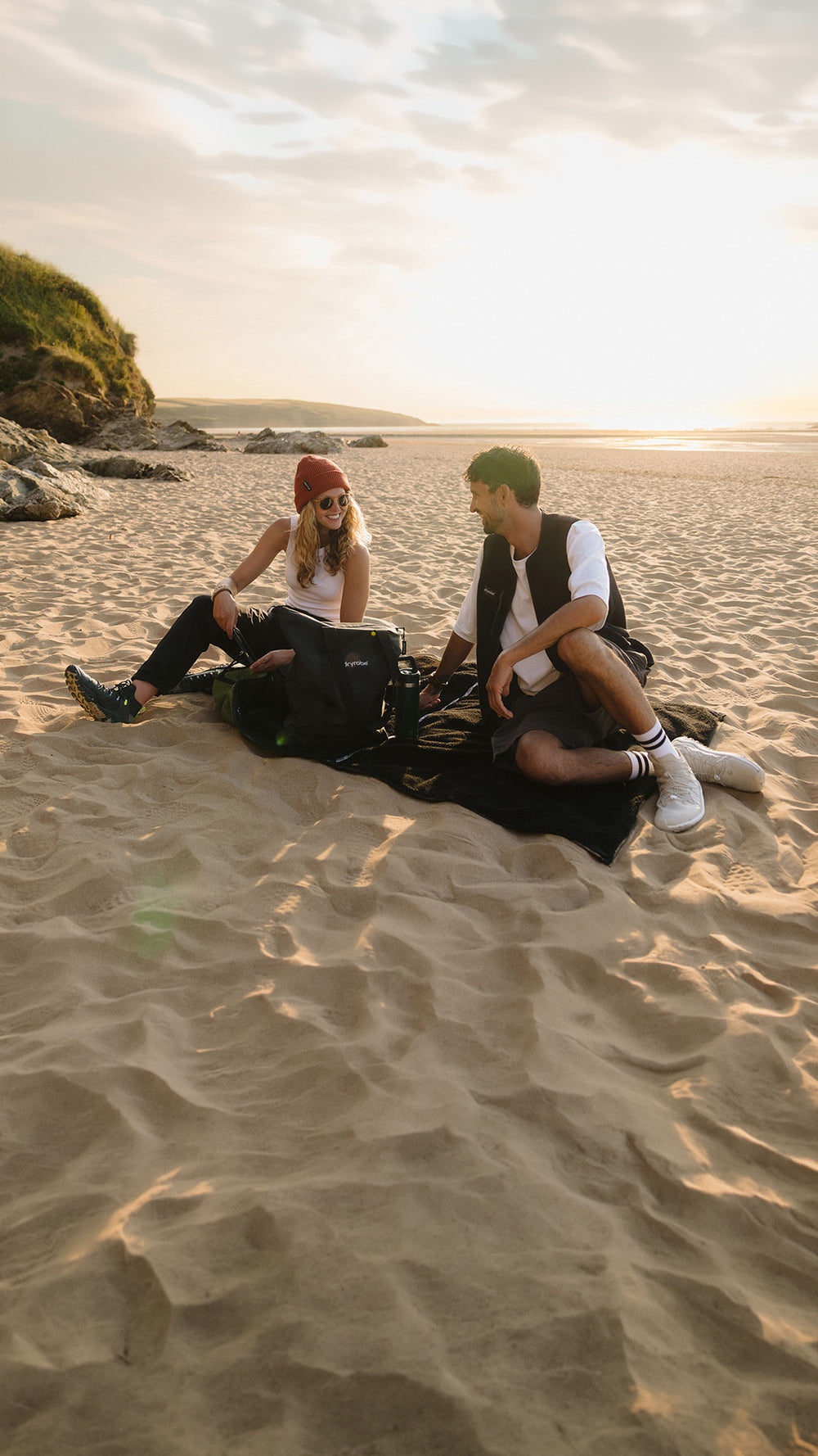 Two people sitting on a black picnic blanket on a sandy beach during golden hour, with the ocean and rocky coastline in the background.