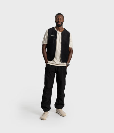 *MALE* A man modeling a black sleeveless vest over a cream t-shirt, black pants, and beige sneakers, standing against a light gray studio background.
