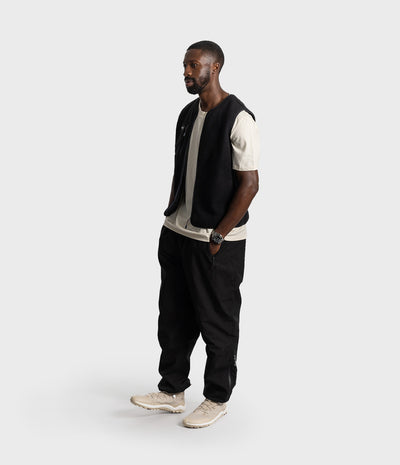 *MALE* A man stands in profile wearing a black sleeveless vest over a cream short-sleeve t-shirt, black pants, and beige sneakers, photographed against a light gray studio background.