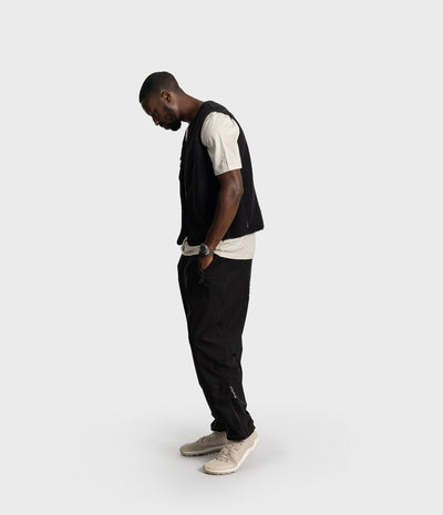 *MALE* A man wearing a black zip-up vest over a cream t-shirt, black pants, and beige sneakers, standing in profile against a light gray studio background.