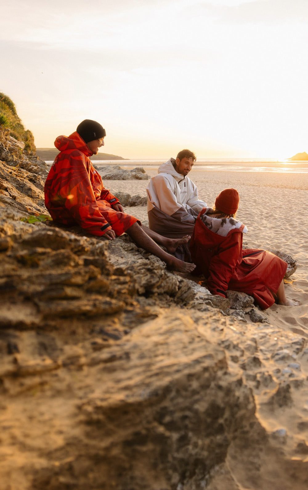 Three people sitting on rocks and sand by the ocean at sunset, wearing Dryrobe Advance.