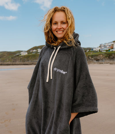 Person wearing a dark grey 'dryrobe' towel robe on a beach with a scenic background.