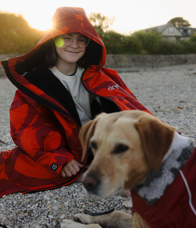 A person wearing a red and orange hooded Dryrobe sits on a gravel surface outdoors at sunset, with a dog in a matching red dog coat in the foreground. The jacket has a black interior lining and a small white logo on the chest.