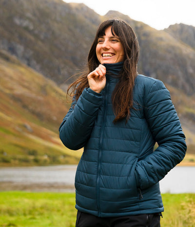 A woman smiling and wearing a dark teal quilted puffer jacket, standing outdoors in front of a mountain and lake background.