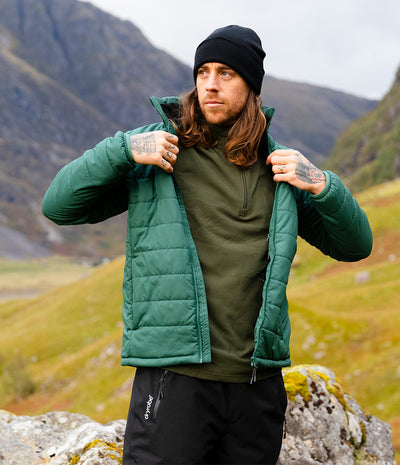 A man wearing a green quilted puffer jacket over an olive green half-zip pullover, black beanie, and black pants, standing outdoors in a mountainous grassy landscape.