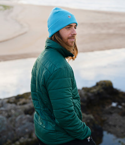 A man wearing a bright blue beanie with a small red circular logo and a dark green quilted puffer jacket, standing outdoors near rocks and water.