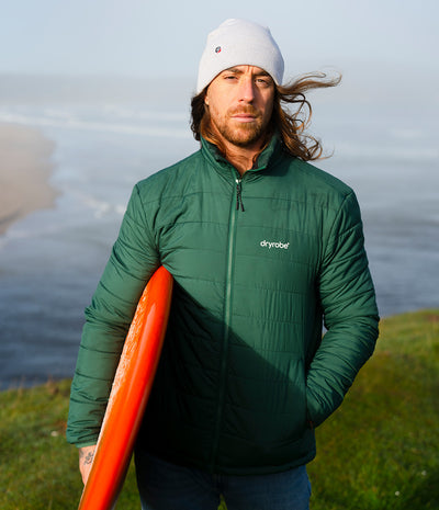 A man wearing a green Dryrobe puffer jacket and a white beanie stands on a grassy cliff overlooking the ocean, holding an orange surfboard.