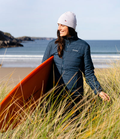 A woman wearing a navy blue puffer jacket and a white beanie walks through beach grass on a sandy dune, holding an orange surfboard with the ocean and coastline in the background.