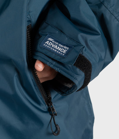 *MALE* Close-up of a dark teal Dryrobe sleeve with a hand sliding into a zippered pocket, showing a black velcro tab and a blue brand patch that reads 'exo-series ADVANCE PERFORMANCE'.