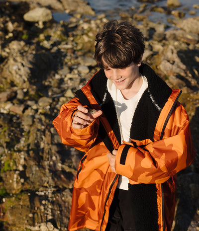 *MALE* A boy wearing a bright orange camouflage-patterned waterproof Dryrobe with black fleece lining, standing outdoors on a rocky shoreline.