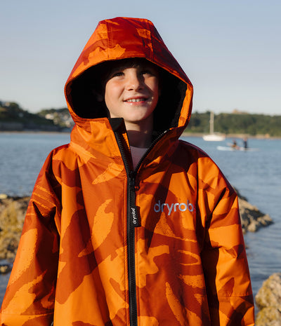 *MALE* A smiling child wearing a bright orange and brown camouflage hooded Dryrobe with a black zipper and the brand name 'dryrob' on the chest, standing outdoors near water and rocks.
