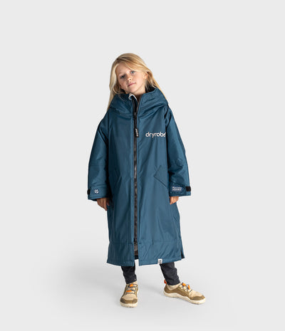 A young girl wearing a long, hooded blue waterproof Dryrobe with a front zipper, standing against a light gray studio background. The coat has the brand name 'dryrobe' printed in white on the left chest.