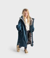 A young girl with blonde hair stands against a light gray studio background wearing a long navy blue hooded Dryrobe with a patterned gray and black lining, a cream t-shirt, dark pants, and gold sneakers.